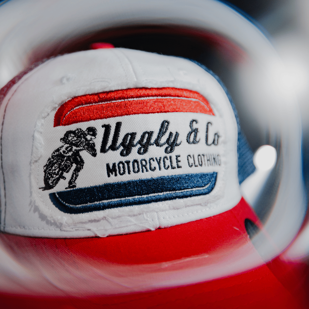 A close up of the front of a red, white and blue flat-track cap with a frayed Uggly&Co badge featuring a motorcycle racing theme