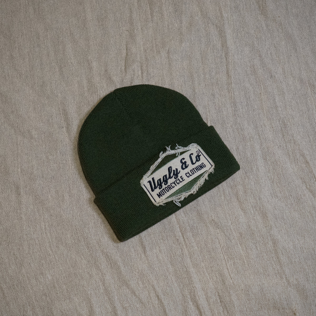 A dark forest green knit beanie with a white embroidered patch reading 'Ugly & Co Motorcycle Clothing' on a light beige fabric background.