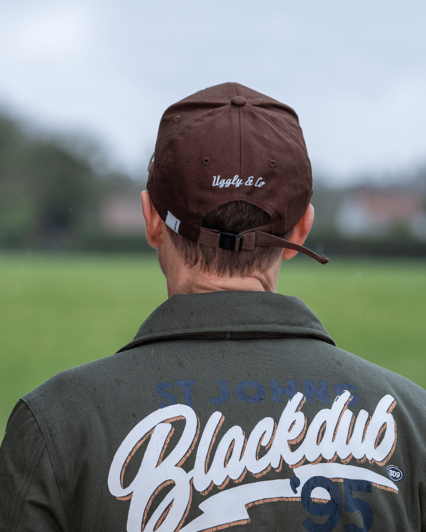 Rear view of a man wearing a brown jacket and trucker hat with 'Uggly & Co' branding, looking at an airplane on a grassy field.