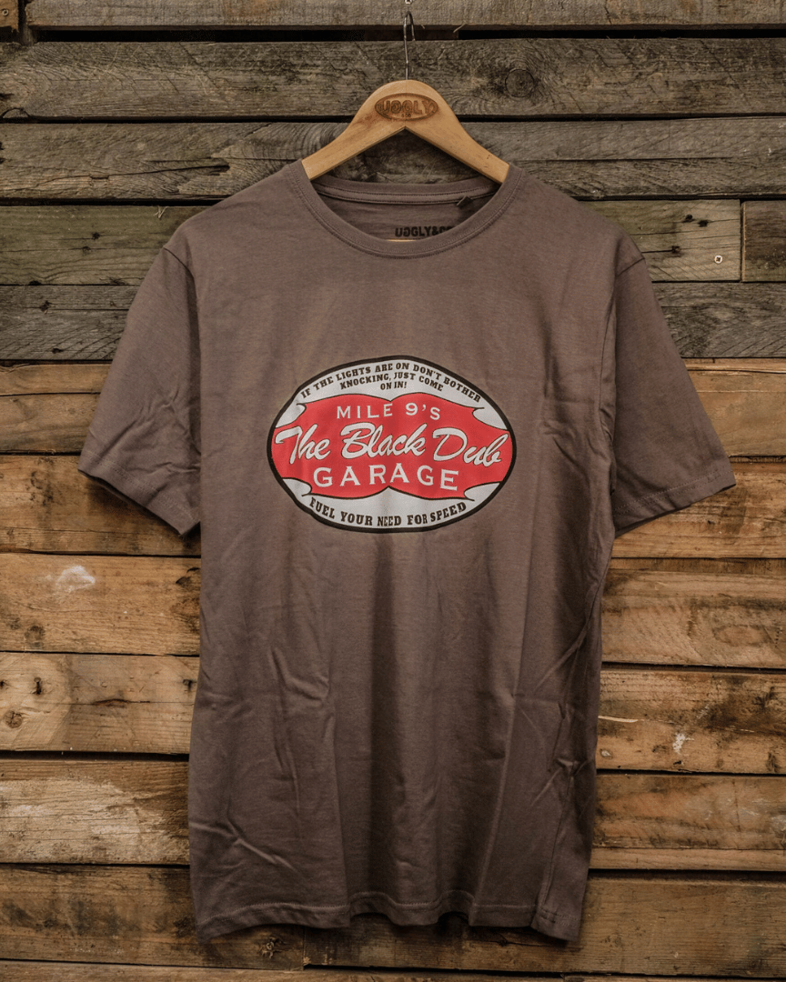 A gray crew neck t-shirt with 'The Black Dub Garage' text printed on the front, displayed against a wooden backdrop.