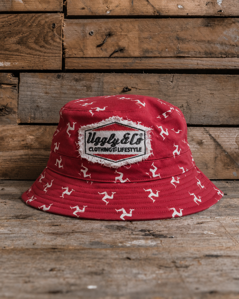 Red bucket hat with white Isle of Man Flag patterns and a Uggly&Co logo on a wooden background