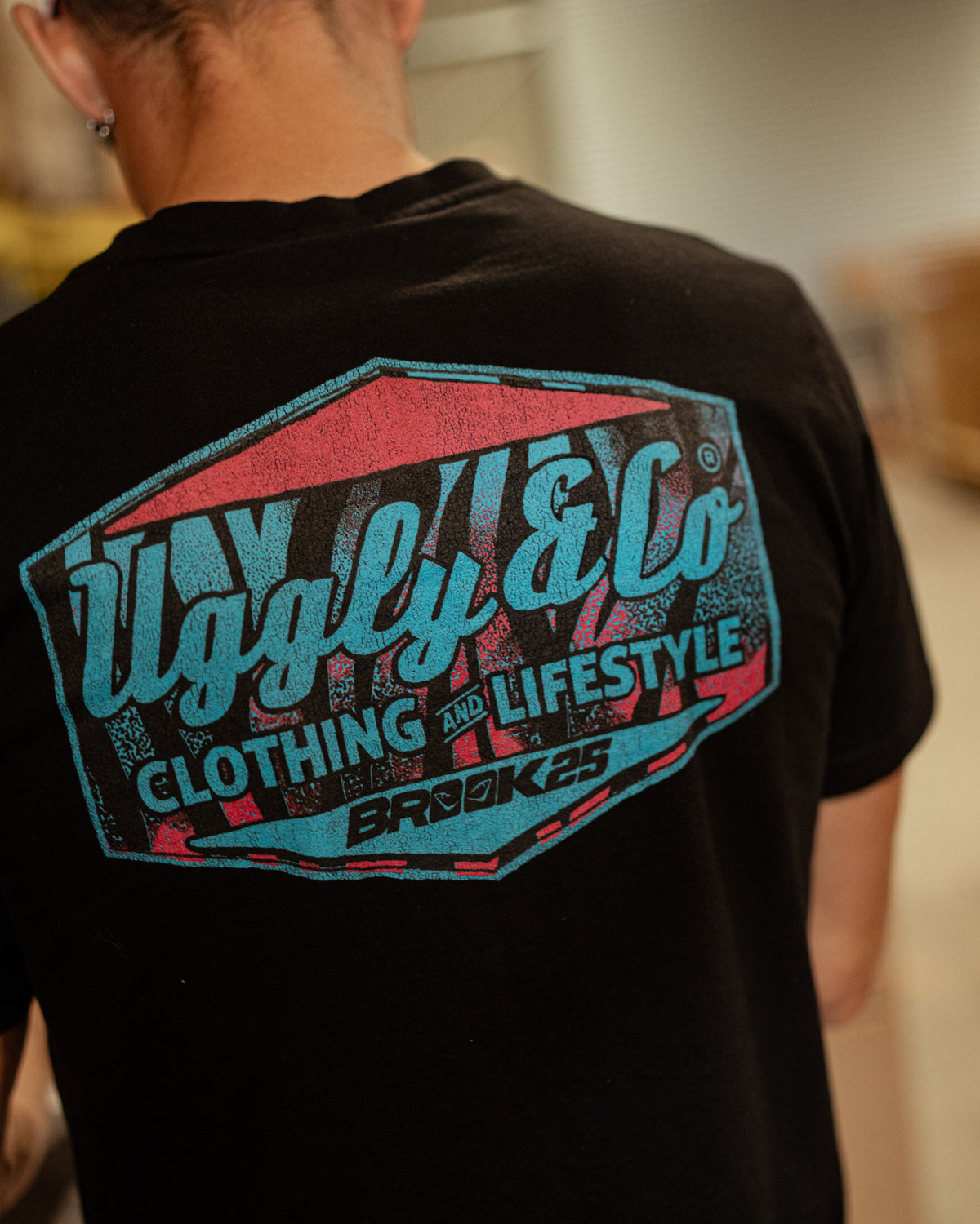 Rear view of a model wearing a Josh Brookes signature diamond black t-shirt with blue and red Uggly&Co diamond logo with a blurred background