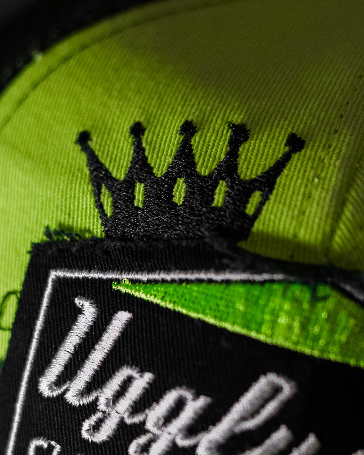 Close-up of the crown logo on the Uggly & Co Fluro Diamond Trucker Hat x Kyle Ryde, photographed against a concrete floor.