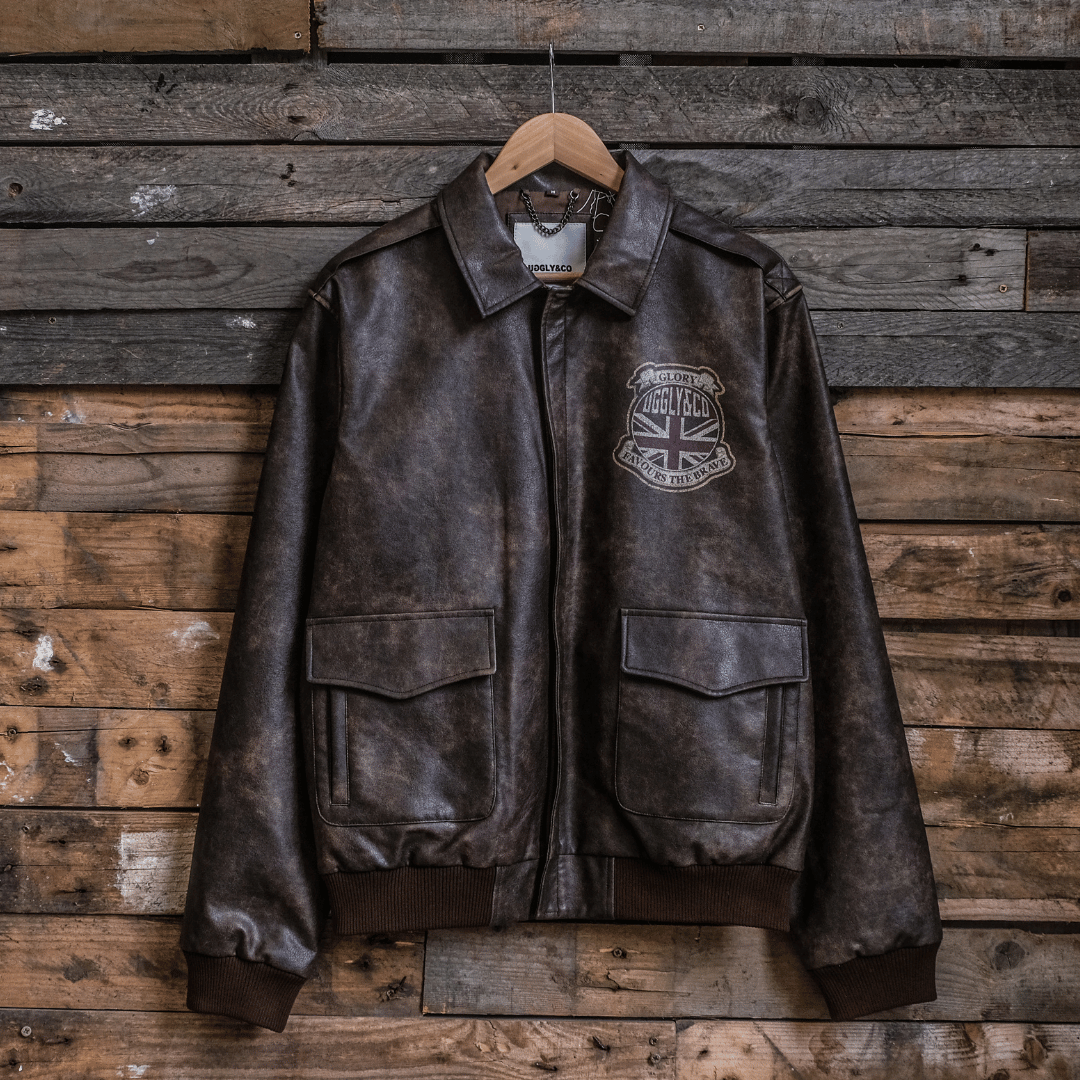 A brown leather aviation bomber jacket with a standing collar, displayed on a wooden hanger against a wooden background.