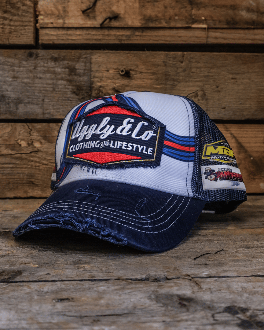 White, red and blue trucker hat with a diamond Uggly&Co logo on the front and other logos showing on the side on a wooden background