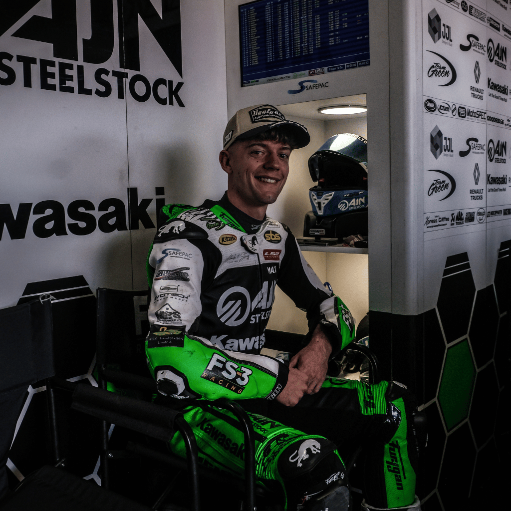 Max Cook in green and black racing leathers, sitting in a garage with branding on the walls and a trucker hat on his head