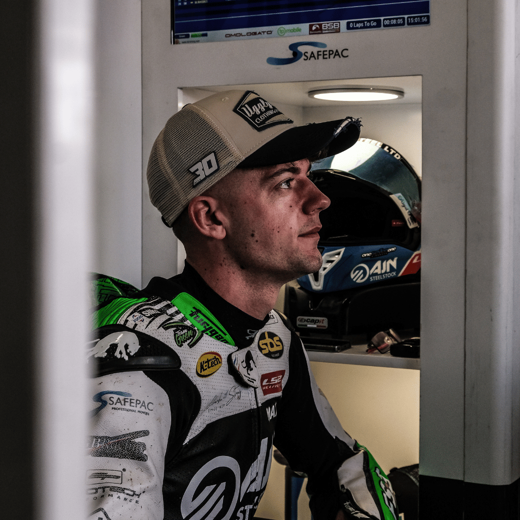 Motorcycle racer Max Cook in a garage wearing his racing leathers and a trucker hat