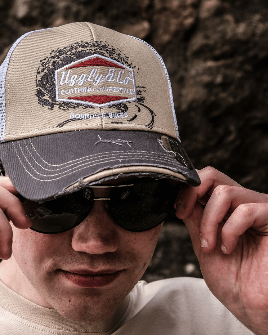 A model putting on sunglasses while wearing a beige and brown trucker cap with a diamond Uggly&Co logo and wave texture design, featuring a white mesh back and a curved peak, with a blurred rocky background.