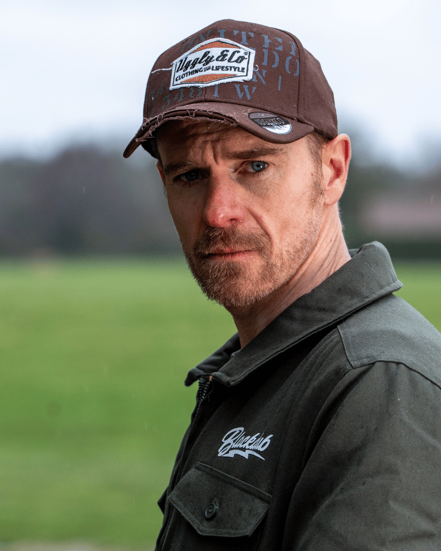 A man looking at the camera while wearing a brown jacket and trucker hat with 'Uggly & Co' branding with a blurred field background