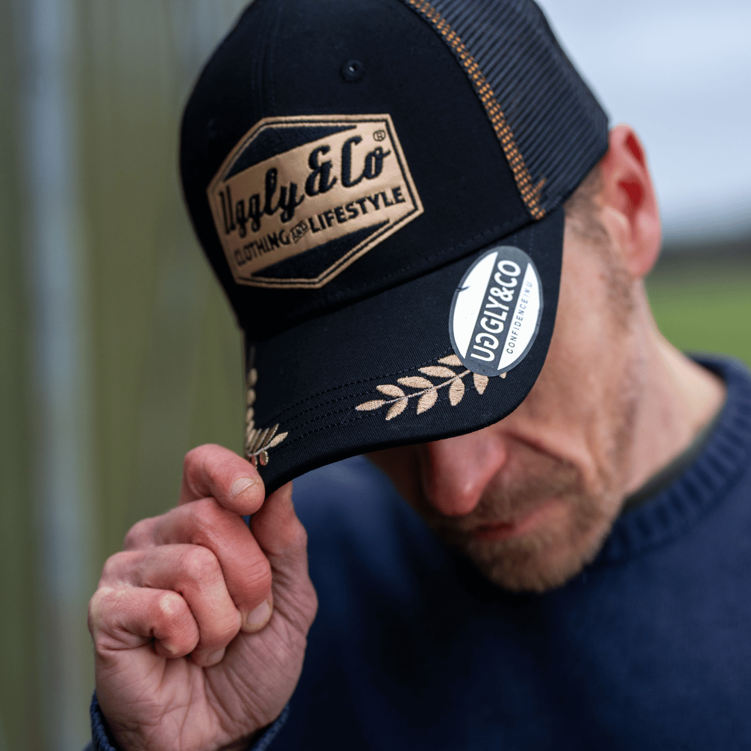 Person wearing a black trucker hat with visible gold branding, holding it with one hand.