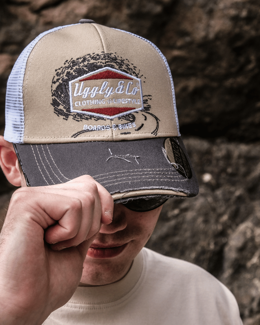 A model looking down while wearing a beige and brown trucker cap with a diamond Uggly&Co logo and wave texture design, featuring a white mesh back and a curved peak, with a blurred rocky background.