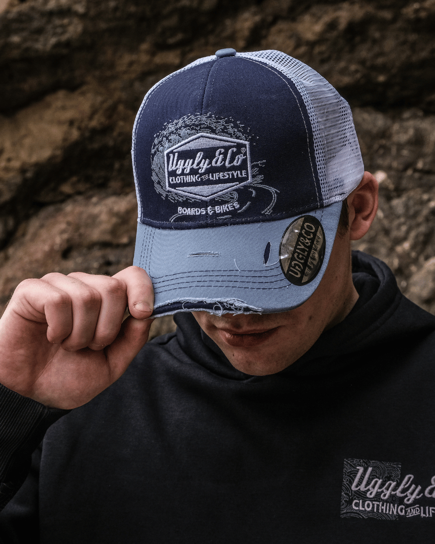A model wearing a blue trucker hat with a white print of the Uggly&Co diamond logo and a light blue wave texture on the front, featuring a navy blue front panel, a sun-faded light blue peak, and a breathable white mesh back, in front of rocks