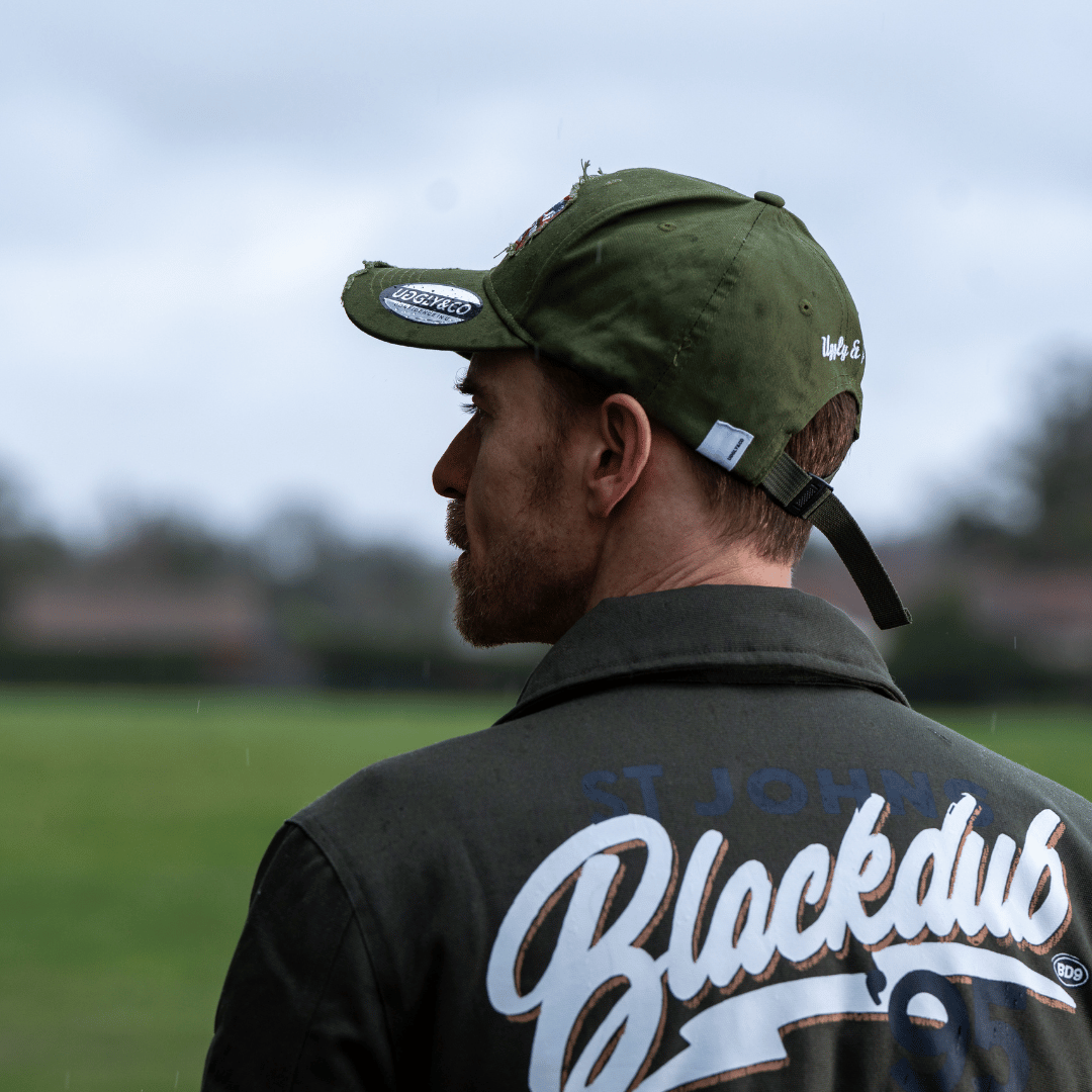 A model side view wearing a green vintage cap with a union jack Uggly&Co brand logo with a blurred background