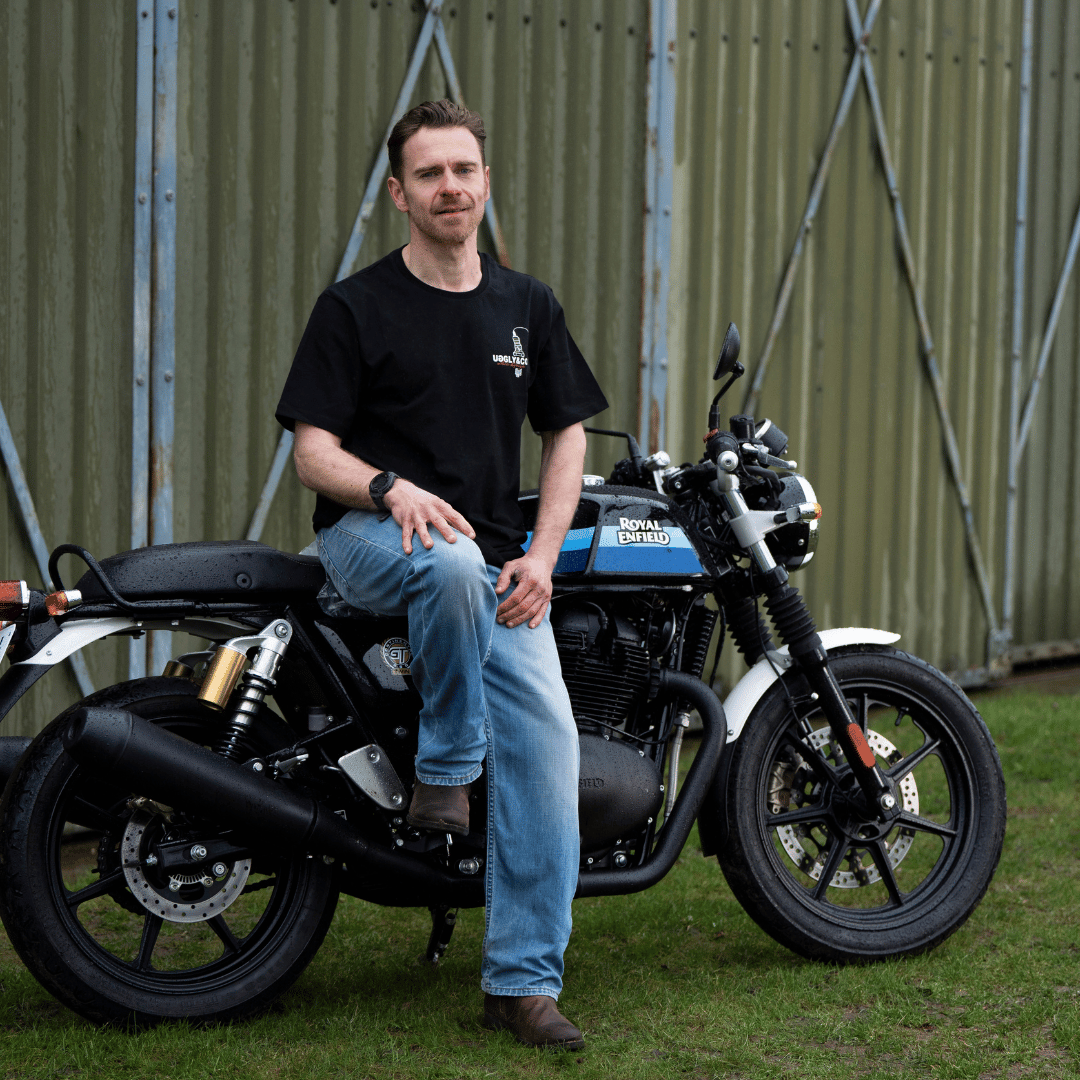 Front view of a model wearing a black t-shirt with a spark plug graphic and the Uggly&Co logo while sitting on a motorcycle