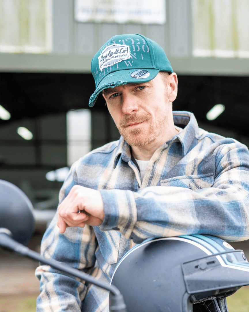 Man wearing a green 'Aviation' Diamond Trucker Hat with distressed black and white Uggly&Co logo and plaid jacket sitting on a motorcycle on an airfield