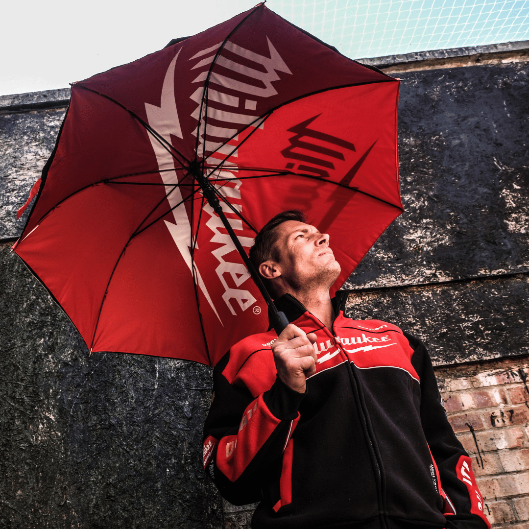 A person holding a red umbrella with Milwaukee Tools and Ducati logos, standing against a wall with a dark color.