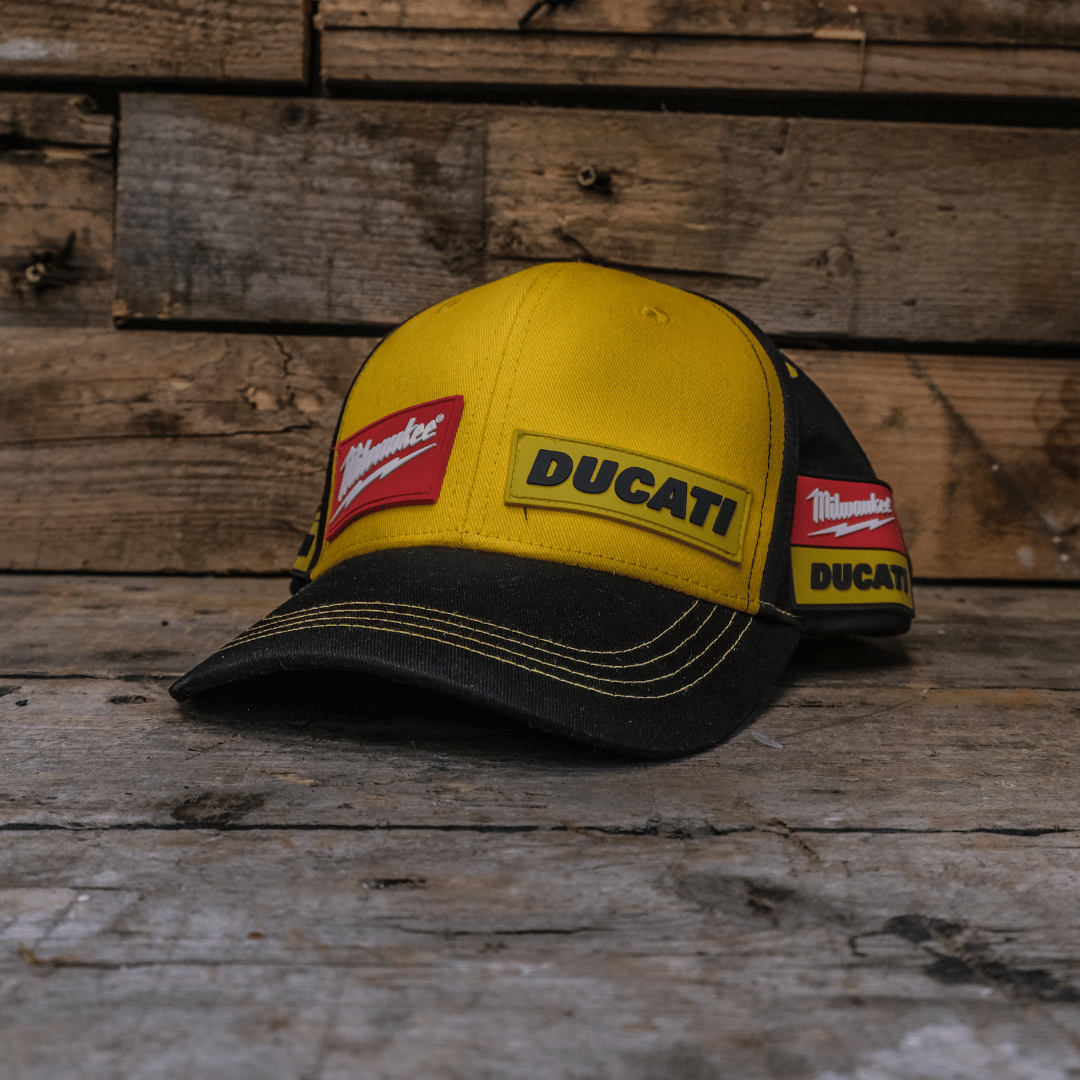 A yellow and black Milwaukee Tools x DUCATI cap with red stitching and embroidered logos placed on a wooden surface.