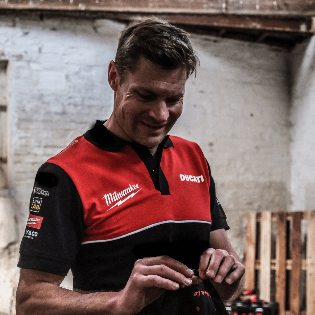 A model looking down and smiling while wearing a Milwaukee x Ducati polo shirt, featuring a black lower half and a red upper torso in a warehouse