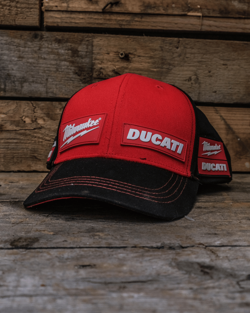 A red and black Milwaukee Tools x DUCATI team-wear cap with embroidered Ducati logo on the front, placed on a wooden surface.