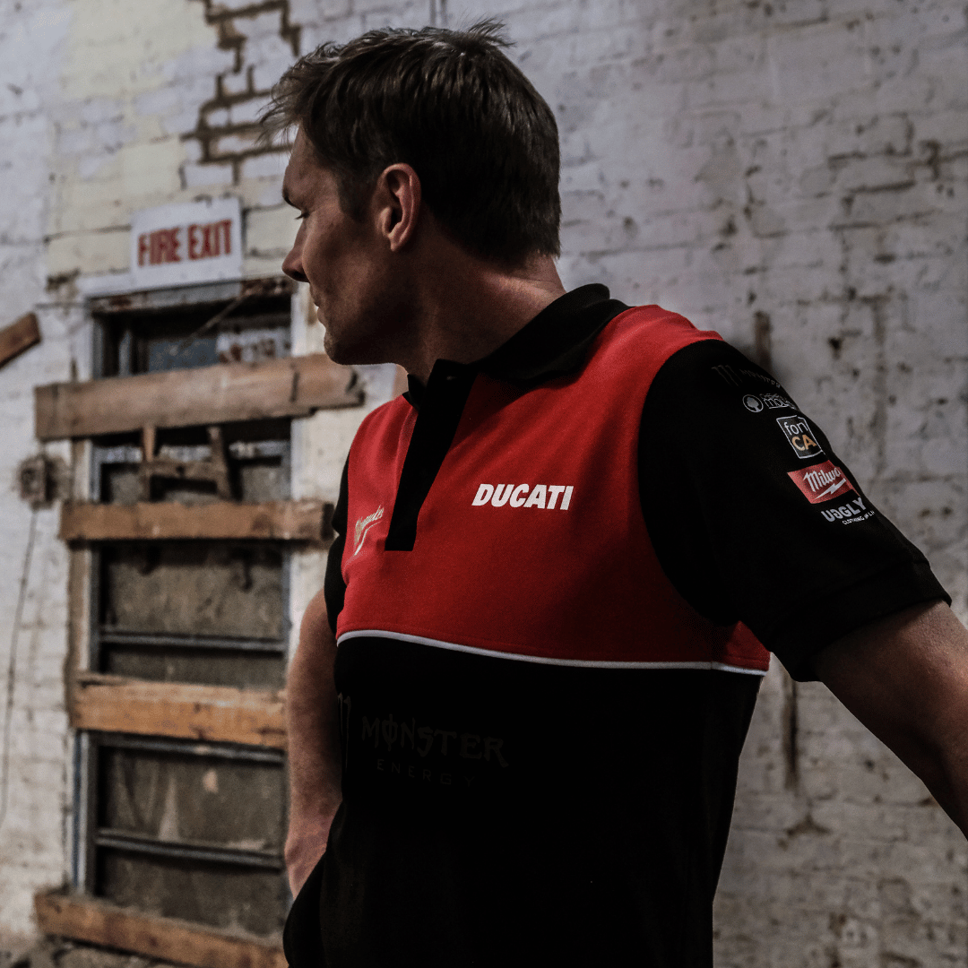 A model looking left while wearing a Milwaukee x Ducati polo shirt, featuring a black lower half and a red upper torso in a warehouse