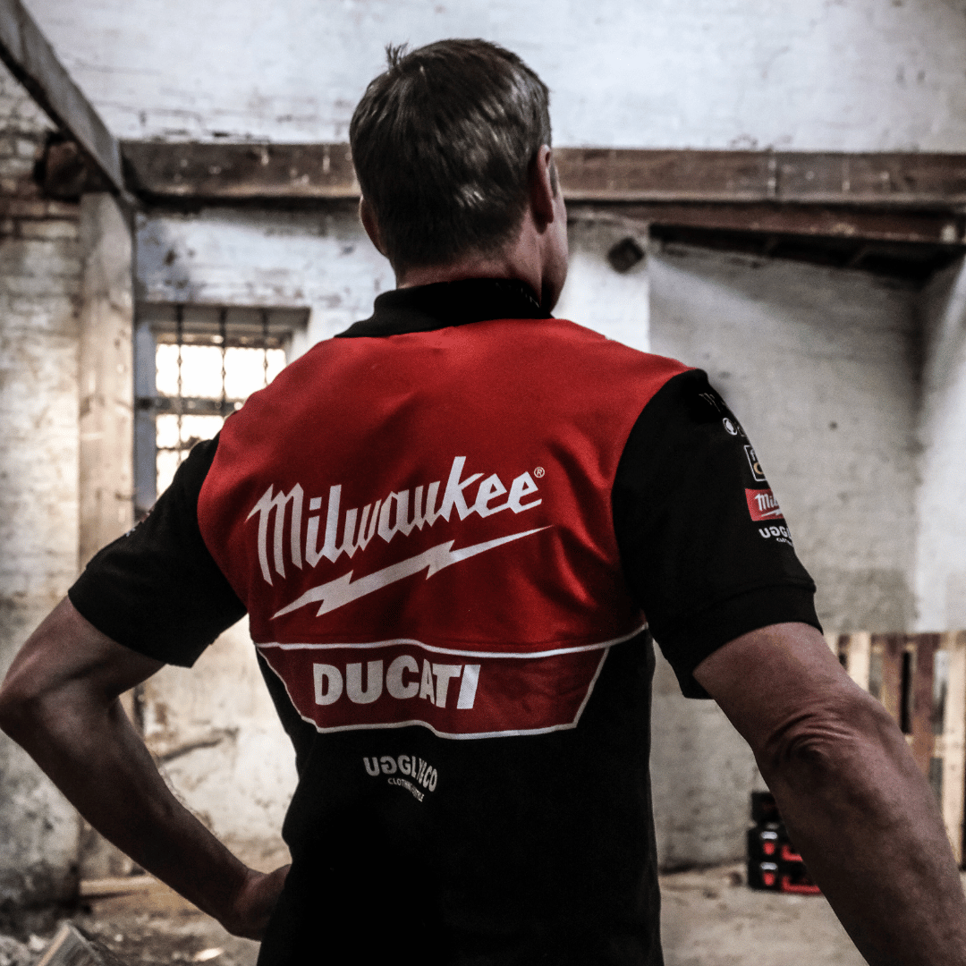 Rear view of a model wearing a Milwaukee x Ducati polo shirt, featuring a black lower half and a red upper torso in a warehouse