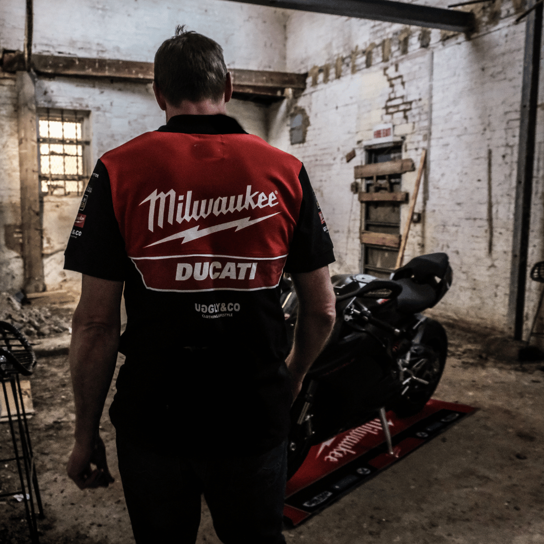 Rear view of a model looking at a motorcycle while wearing a Milwaukee x Ducati polo shirt, featuring a black lower half and a red upper torso in a warehouse