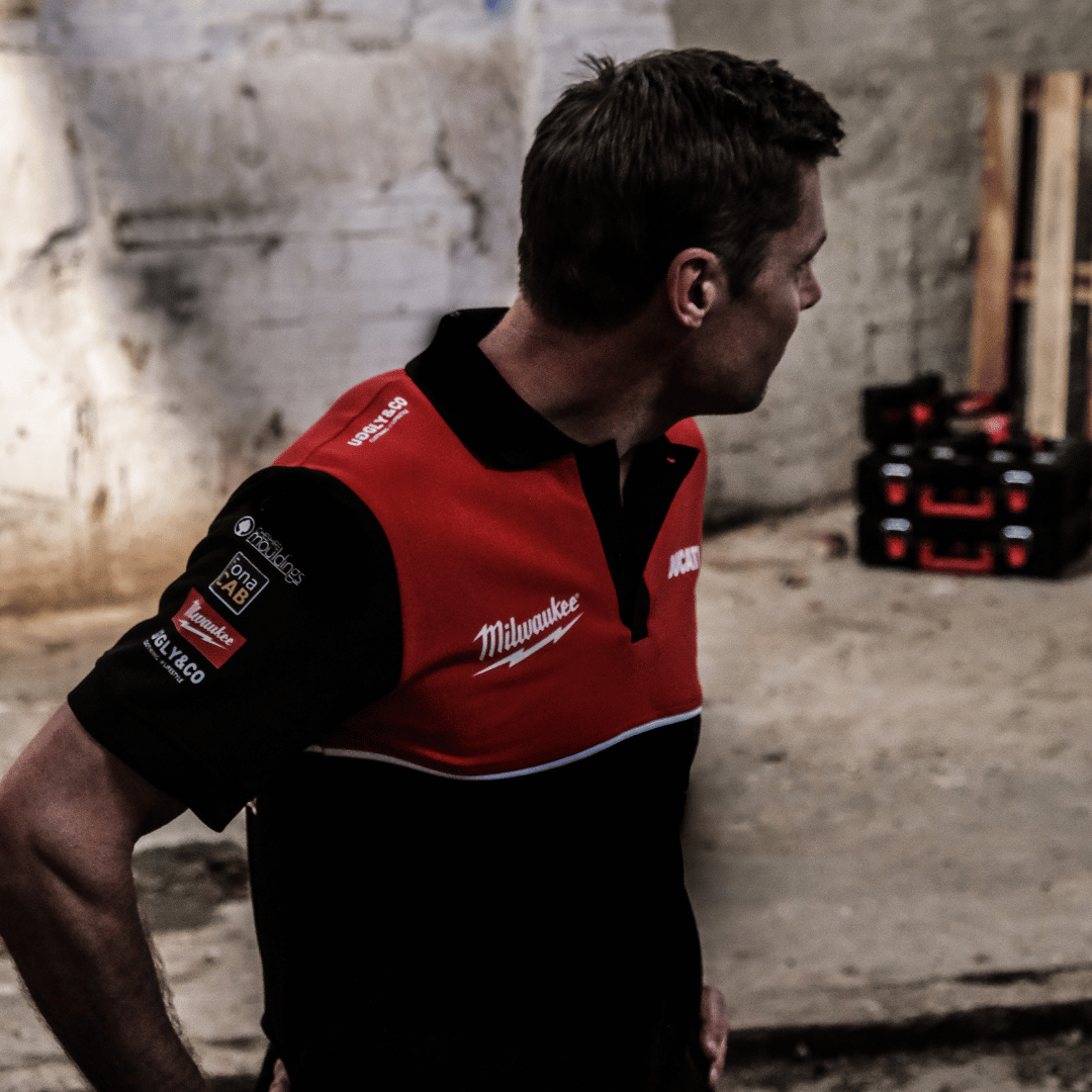 A model looking right while wearing a Milwaukee x Ducati polo shirt, featuring a black lower half and a red upper torso in a warehouse