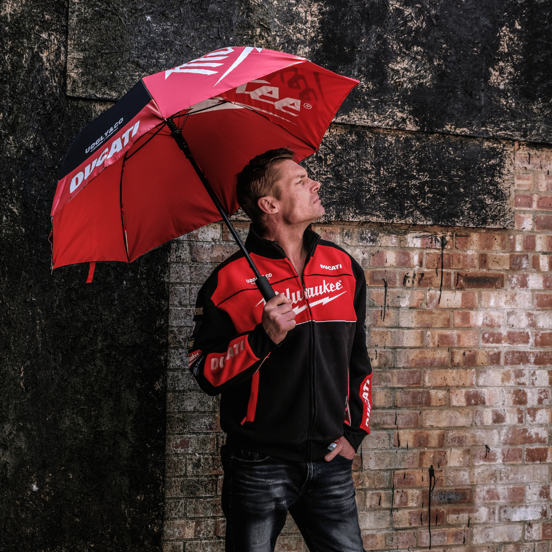 A model holding a red and black Milwaukee x Ducati umbrella with Milwaukee logo against a brick wall backdrop