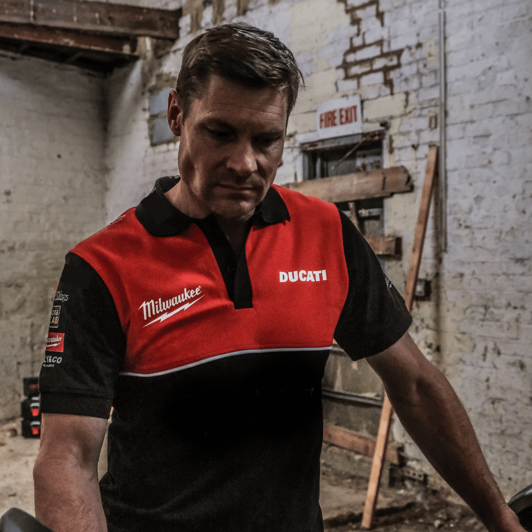 A model looking down at a motorcycle out of shot while wearing a Milwaukee x Ducati polo shirt, featuring a black lower half and a red upper torso in a warehouse