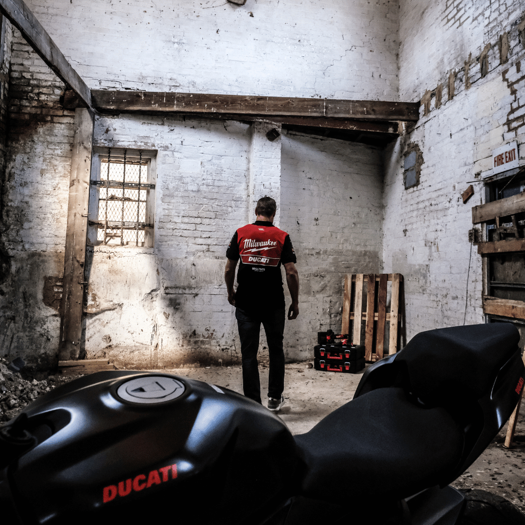 A model walking away from the camera while wearing a Milwaukee x Ducati polo shirt, featuring a black lower half and a red upper torso in a warehouse
