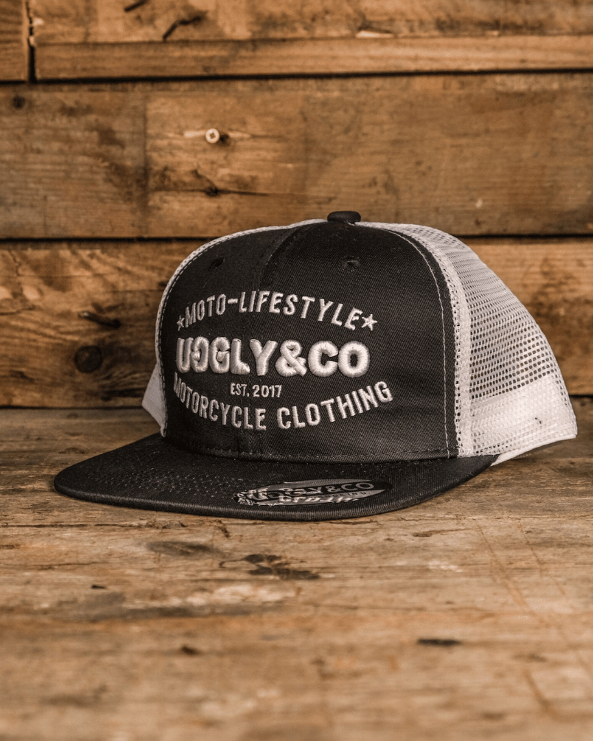 A black and white trucker cap with 'Moto-Lifestyle' text and the 'UGGLY&CO' brand logo on it, placed on a wooden surface.