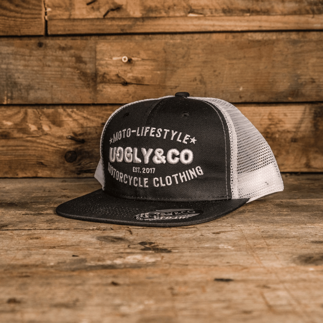 A black and white trucker cap with 'Moto-Lifestyle' text and the 'UGGLY&CO' brand logo on it, placed on a wooden surface.