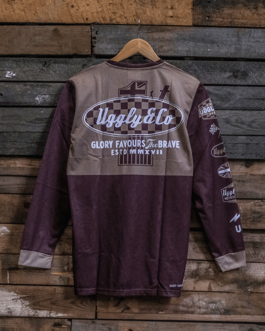 A long-sleeved brown and beige motocross jersey with checkered patterns and text graphics hanging on a wooden background backwards.