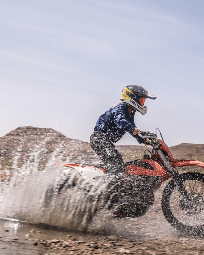 Riding a motocross bike through a puddle in the desert in a blue Uggly&Co motocross jersey