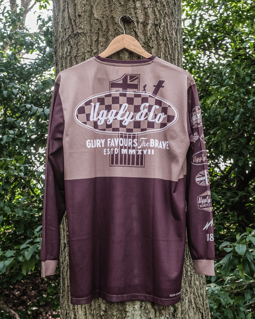 A long-sleeved brown and beige motocross jersey with checkered patterns and text graphics hanging on a tree backwards.