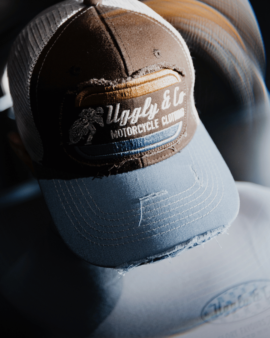 Top view of a dusty blue trucker hat with a brown front panel, featuring a vintage flat track Uggly&Co logo with a blurred background