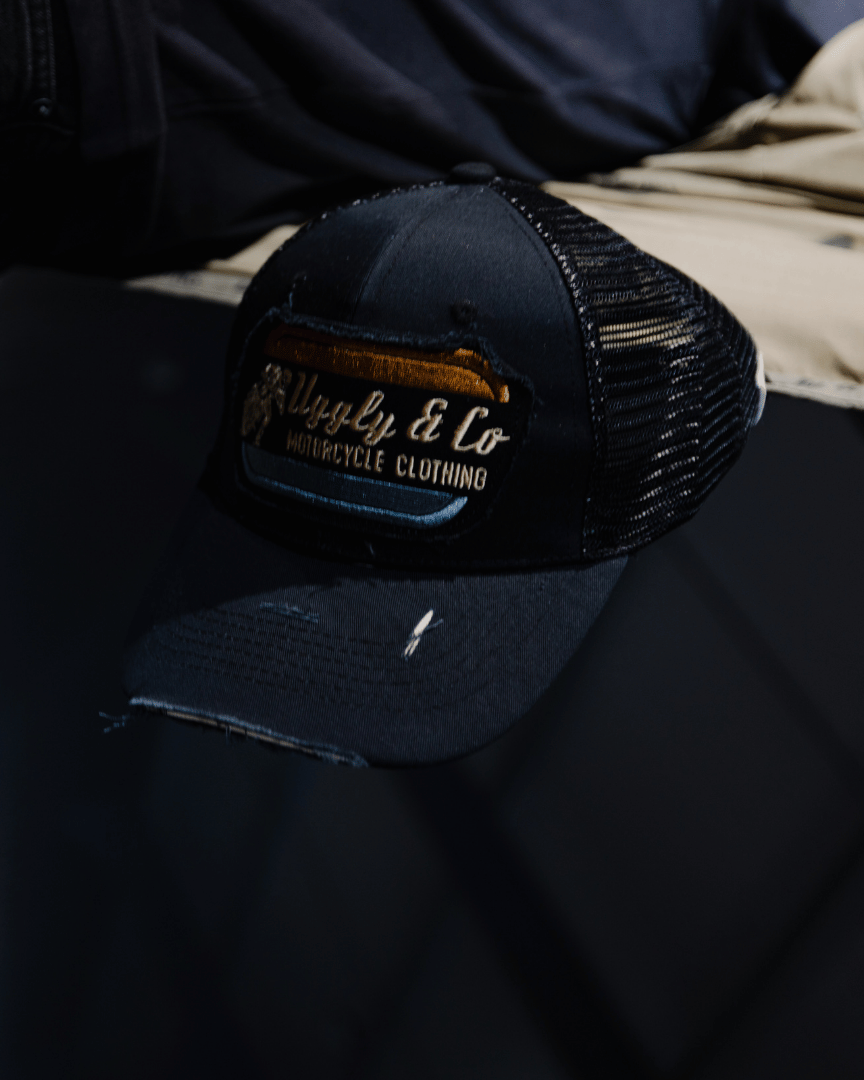 An all-black trucker hat with a vintage-inspired flat track racing logo in yellow and blue on the front on a blurred background