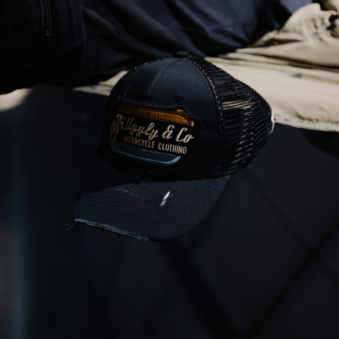An all-black trucker hat with a vintage-inspired flat track racing logo in yellow and blue on the front on a blurred background