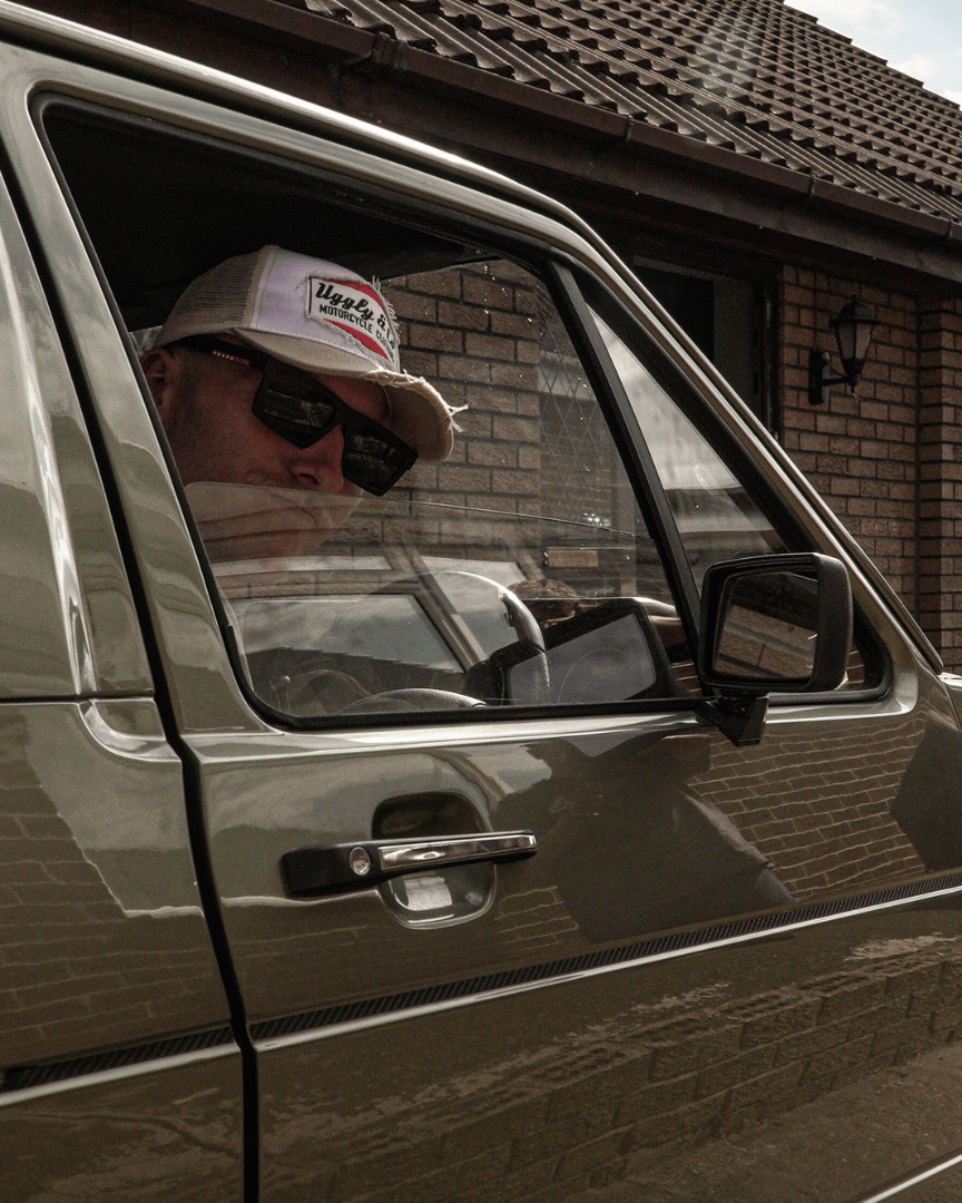 A model looking out of a car window while wearing a beige and white Uggly & Co motorcycle clothing trucker hat with a mesh back and an embroidered logo on the front