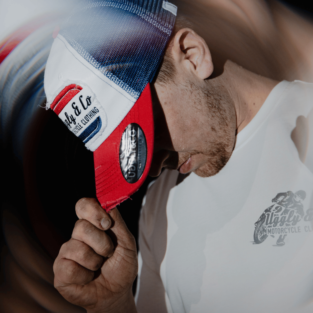 A model looking down while wearing a red, white and blue flat-track cap with a frayed Uggly&Co badge featuring a motorcycle racing theme with a spinning blurred background