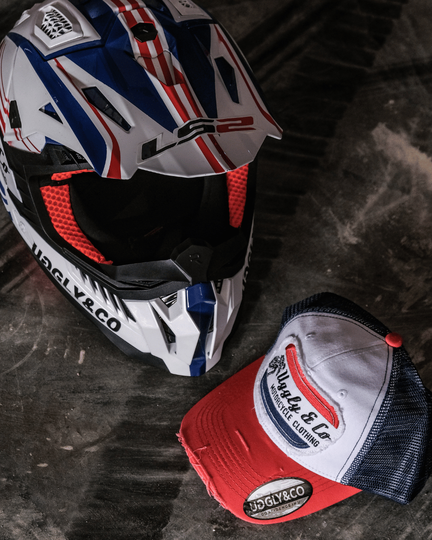 A red, white and blue flat-track cap with a frayed Uggly&Co badge featuring a motorcycle racing theme, on the floor with a matching motocross helmet