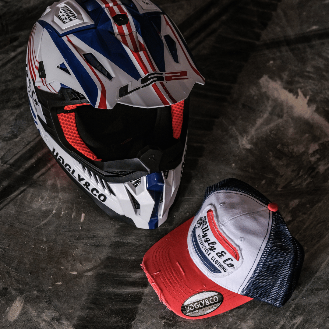 A red, white and blue flat-track cap with a frayed Uggly&Co badge featuring a motorcycle racing theme, on the floor with a matching motocross helmet