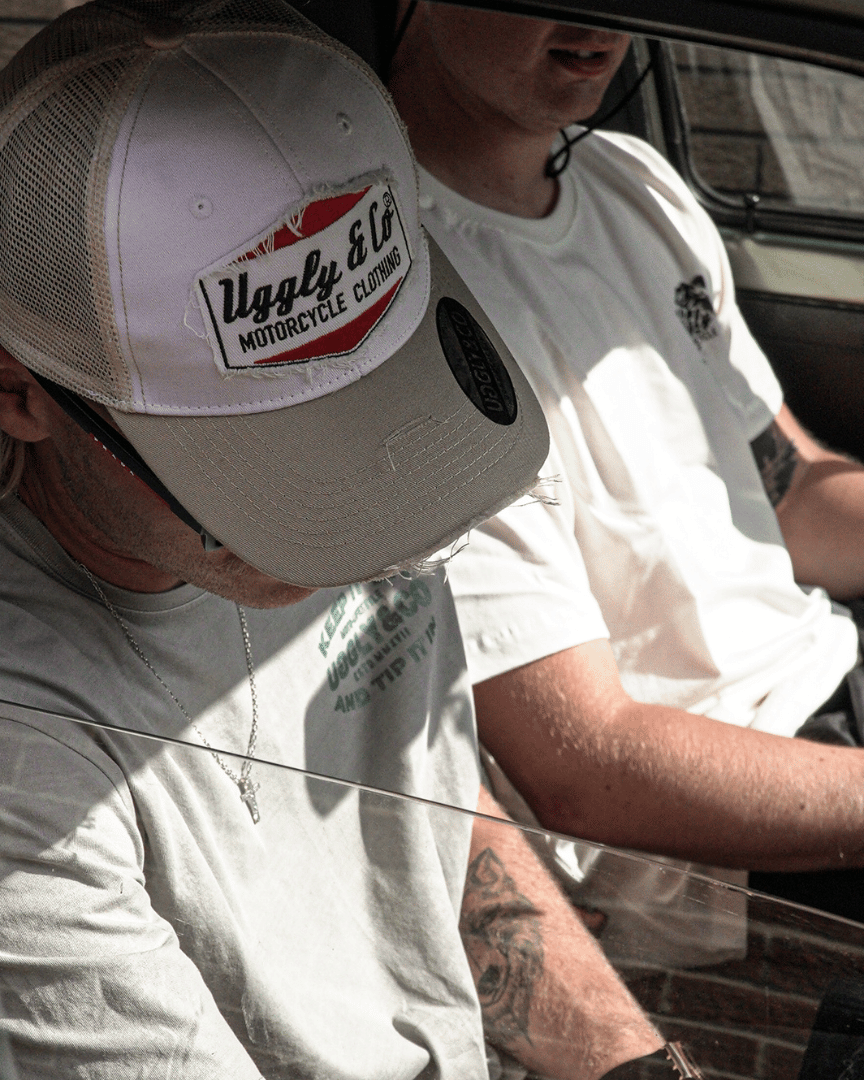 A model looking down while sitting in a car, wearing a beige and white Uggly & Co motorcycle clothing trucker hat with a mesh back and an embroidered logo on the front