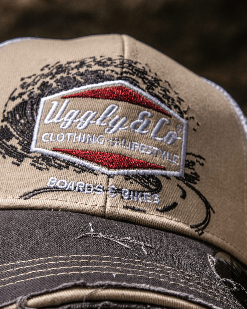 Close up of a beige and brown trucker cap with a diamond Uggly&Co logo and wave texture design, featuring a white mesh back and a curved peak, with a blurred background.