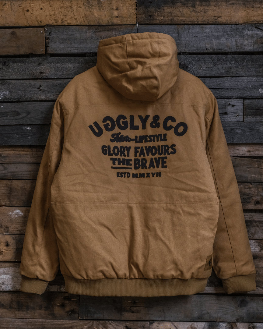A brown U&C chore jacket with a hood, featuring the Uggly&Co script logo and the 'Glory Favours the Brave' moto lifestyle statement on the back, displayed on a wooden background.