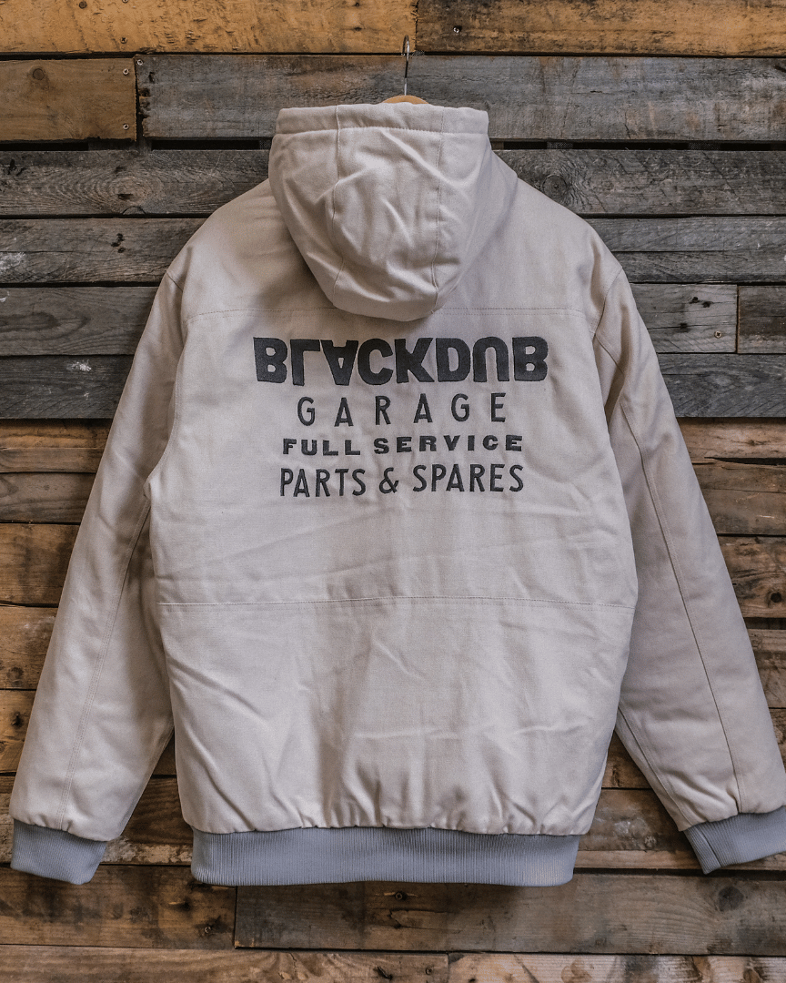 A beige-colored chore jacket with 'BRICKDUB GARAGE PARTS & SPARES' logo on the back, displayed against a wooden background.