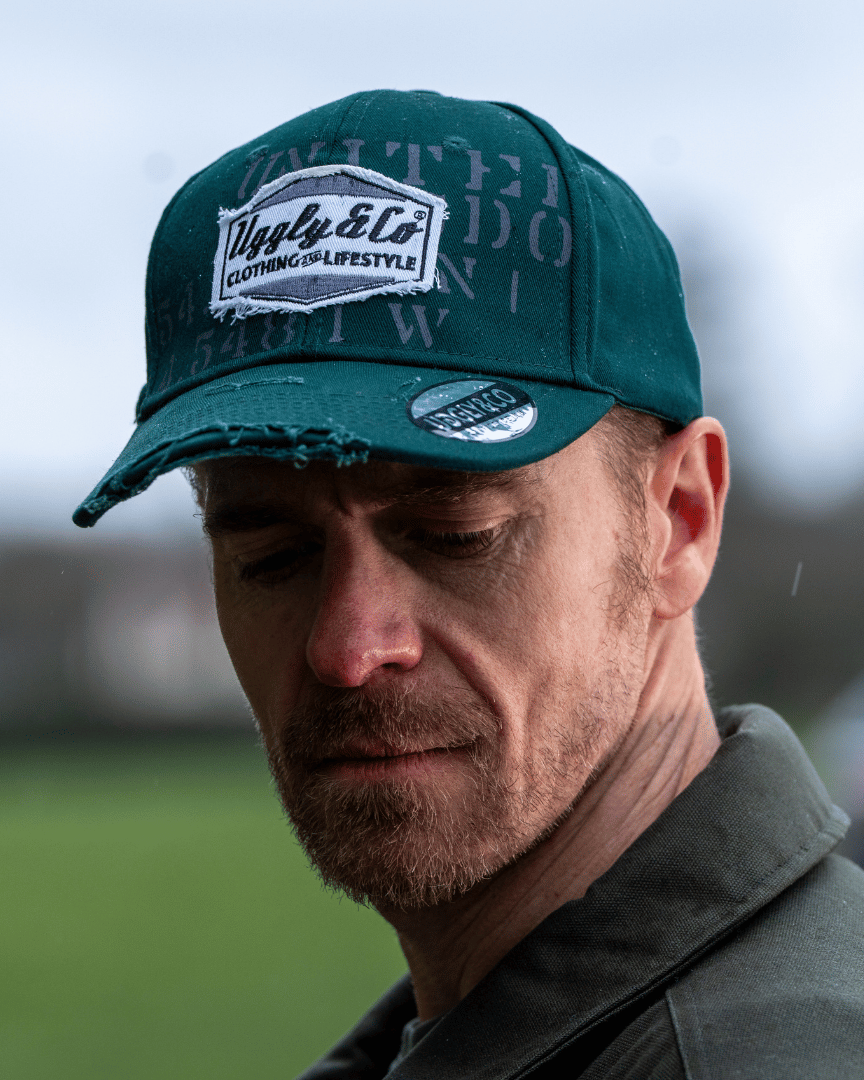 Close up of a man wearing a green 'Aviation' Diamond Trucker Hat with distressed black and white Uggly&Co logo on an airfield
