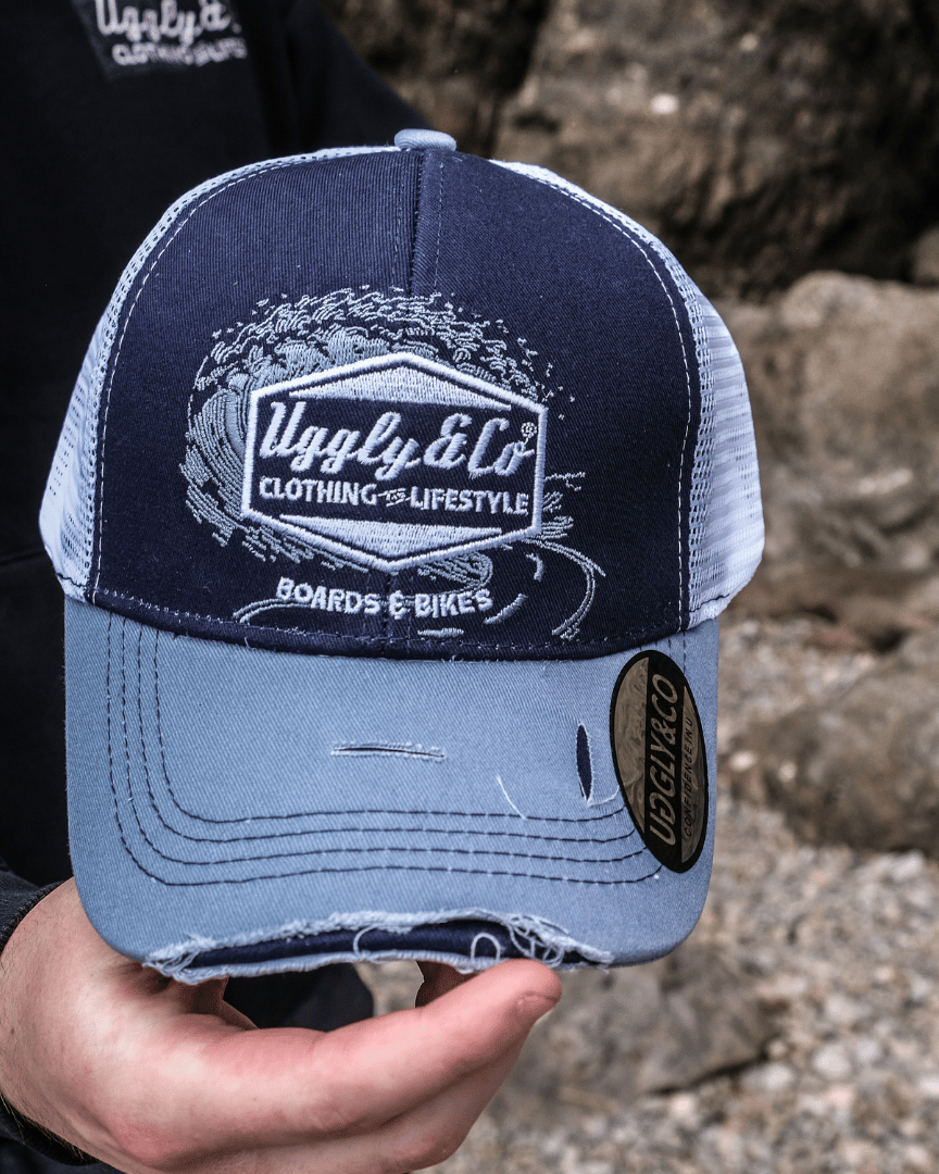 A model holding a blue trucker hat with a white print of the Uggly&Co diamond logo and a light blue wave texture on the front, featuring a navy blue front panel, a sun-faded light blue peak, and a breathable white mesh back, with rocks in the background