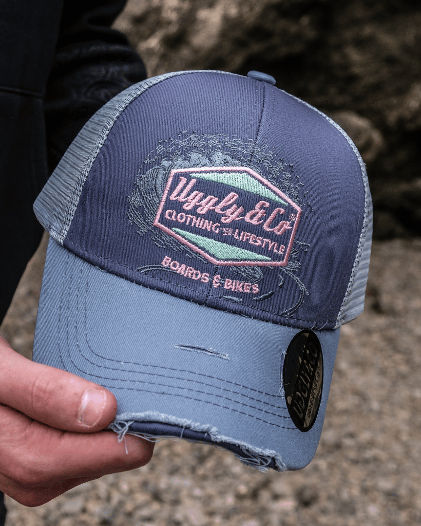 A model holding a trucker hat with a purple front panel and a blue mesh back, featuring a pink diamond Uggly&Co logo and the text 'Boards & Bikes' with a blurred rocky background