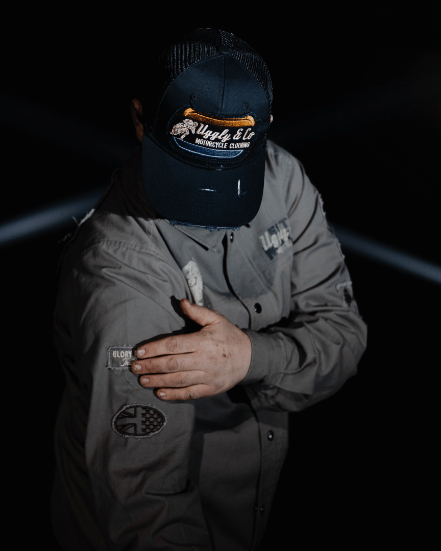 A model wearing an all-black trucker hat with a vintage-inspired flat track racing logo in yellow and blue on the front at night